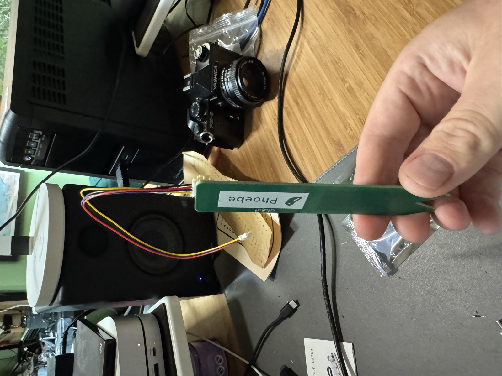Hand holding the green Prong soil sensor board with the Phoebe logo on the silkscreen and bodge wires soldered to the connector